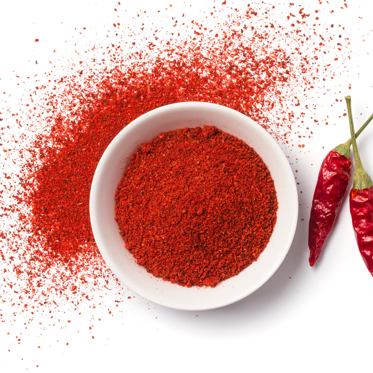 Ground red chili powder scattered around a white bowl on white surface
