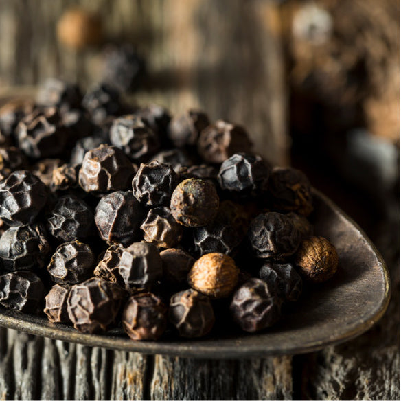 Whole black peppercorns on a metal spoon against a rustic wooden surface
